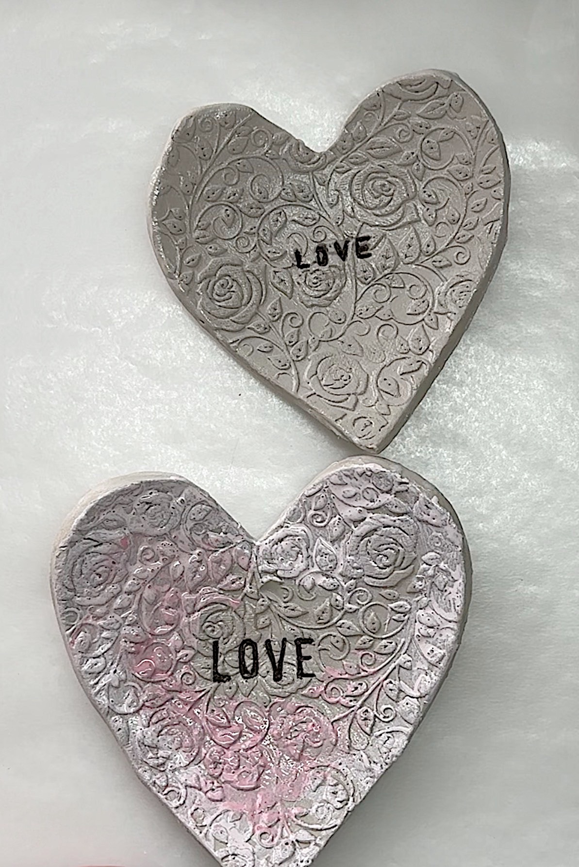 Two heart-shaped clay decorations with embossed floral patterns and the word "LOVE" in black letters, placed on a white surface.