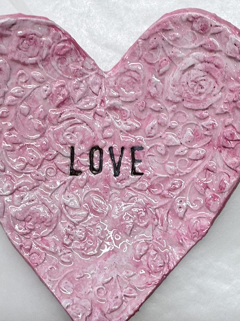 Pink heart-shaped ceramic plate with raised floral patterns and the word "LOVE" in black letters at the center, placed on a white background.