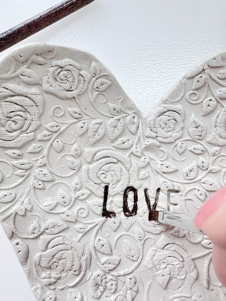 A hand uses a tool to carve the word "LOVE" into a heart-shaped piece of embossed clay with a floral pattern.