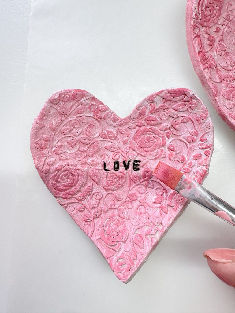 A hand-painted pink heart-shaped clay ornament with floral patterns and the word "LOVE" in black letters, being brushed with paint.