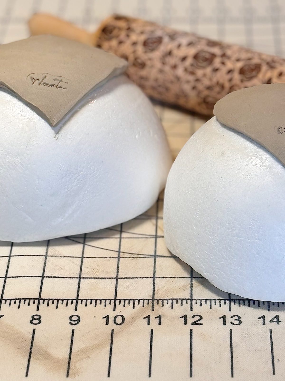Two white foam half-spheres with gray clay pieces on top sit on a gridded work surface; a patterned rolling pin is in the background.