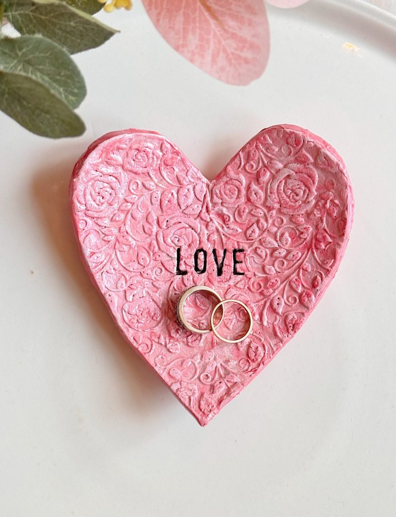A pink textured heart-shaped dish with the word "LOVE" and two gold rings placed on it, set on a white surface with green leaves and pink flower petals nearby.