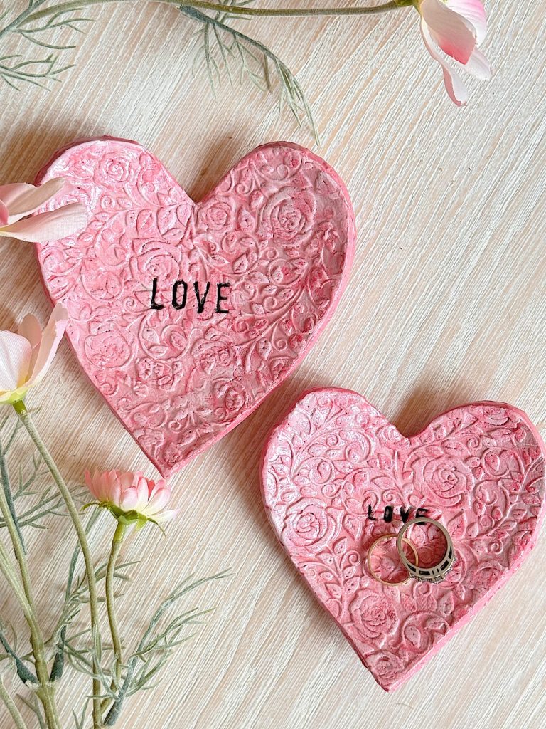 Two pink, heart-shaped ceramic dishes with embossed floral patterns and the word "LOVE"; one dish holds two silver rings, surrounded by light pink flowers on a wooden surface.