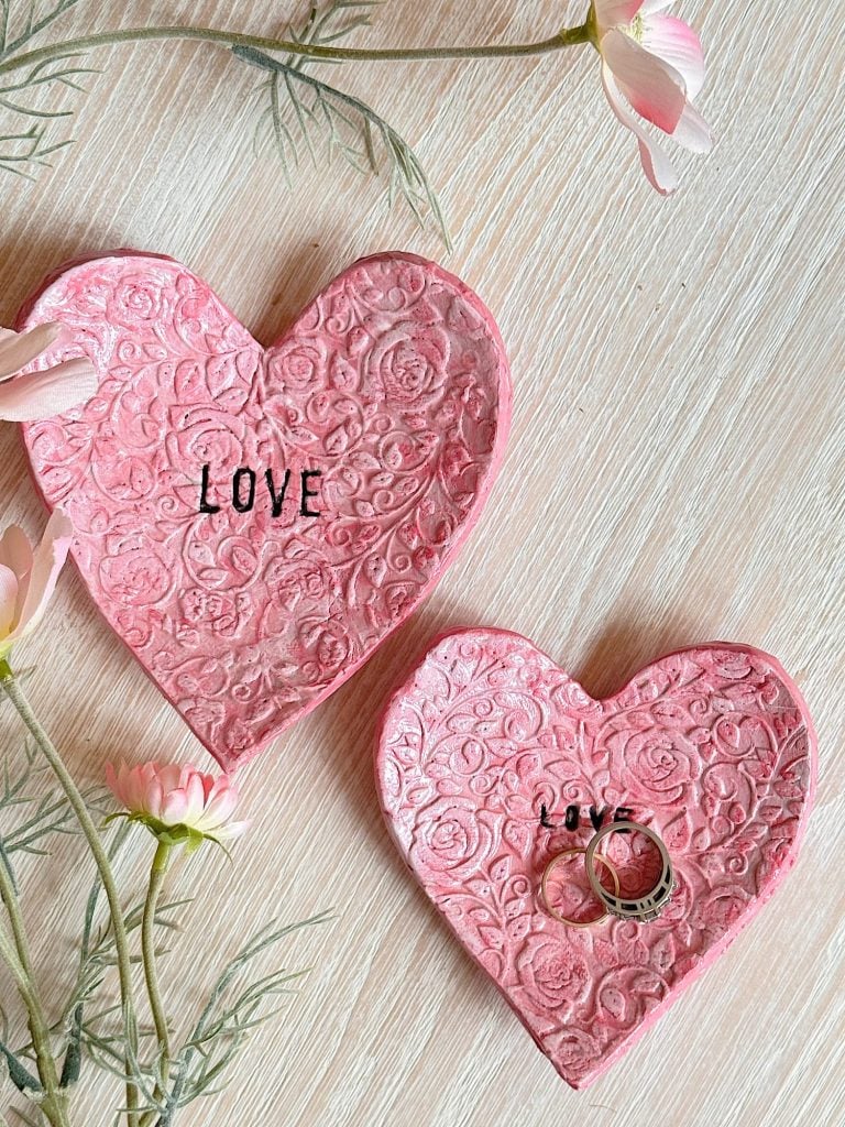Two pink, heart-shaped ceramic dishes with embossed floral patterns and the word "LOVE"; one holds two rings, surrounded by pink flowers on a light wooden surface.