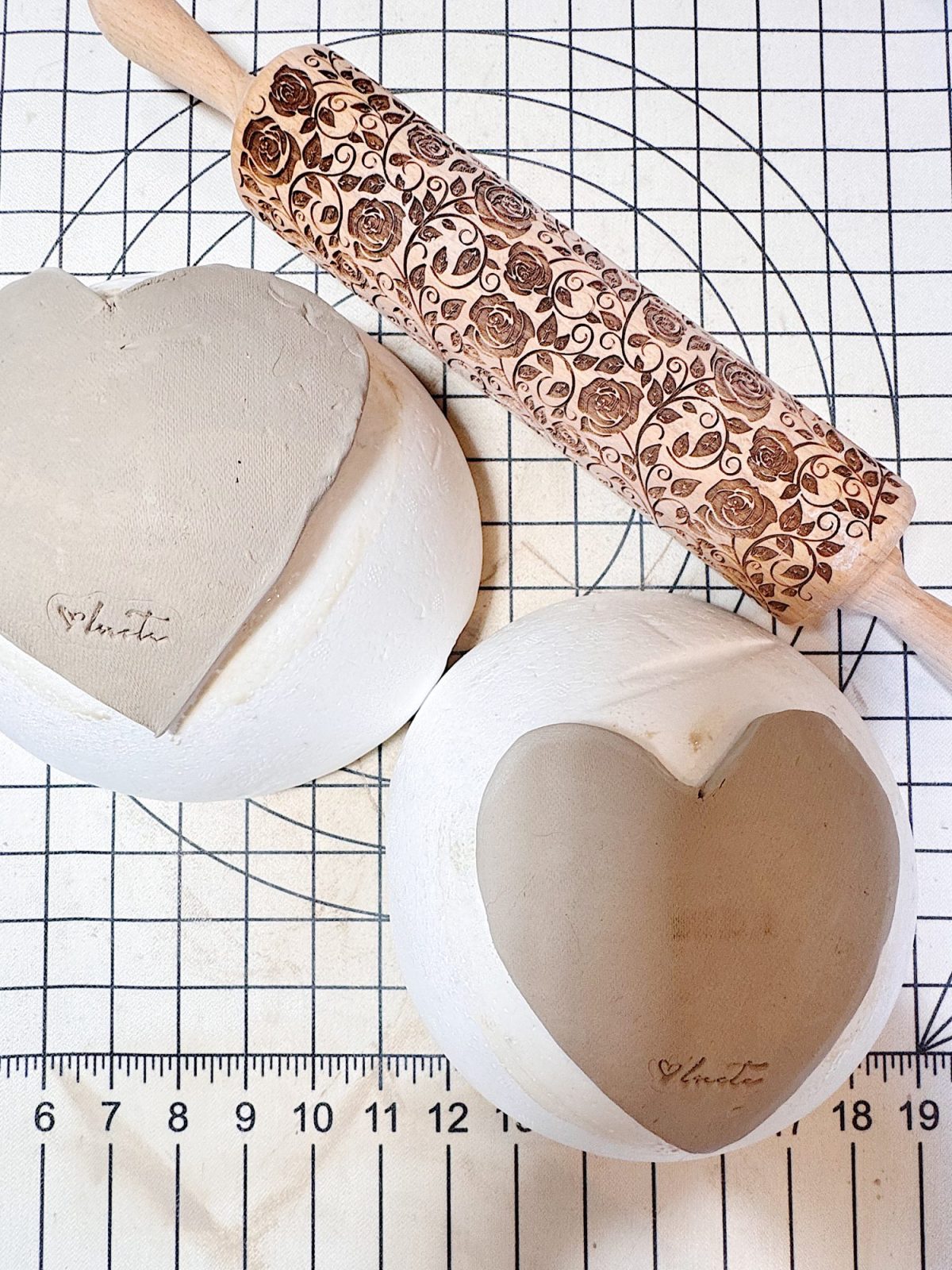 A patterned rolling pin, a heart-shaped slab of clay, and a heart-shaped clay dish with "heart" inscribed, all placed on a gridded work surface.