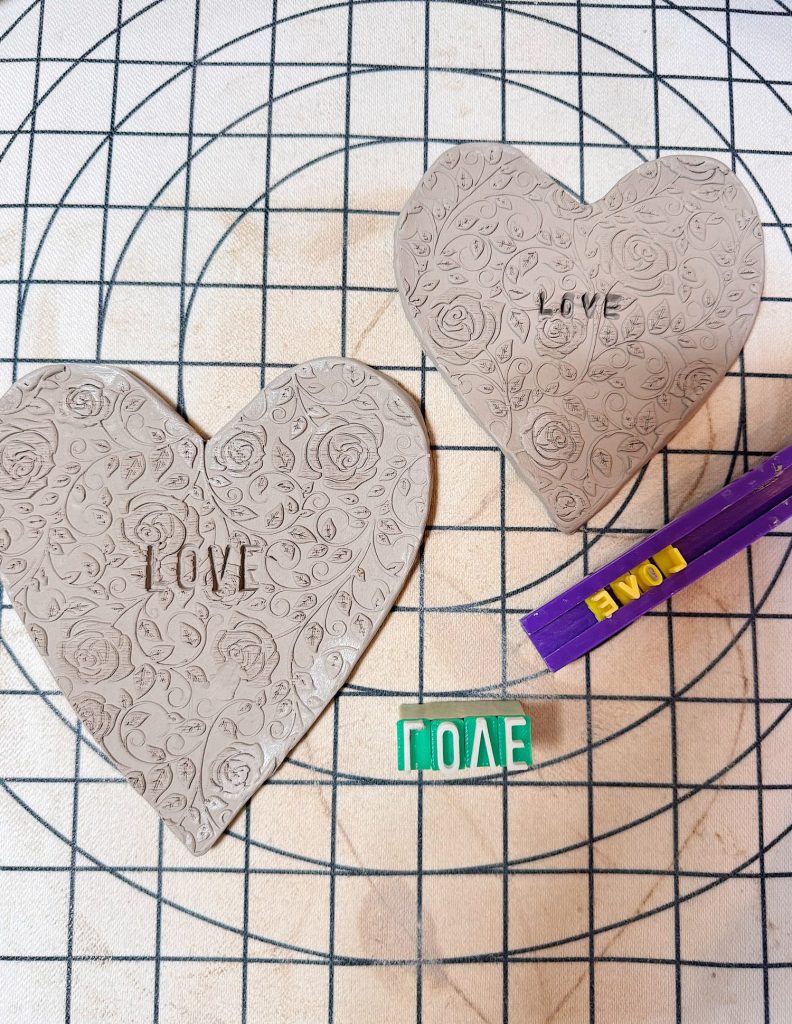Two heart-shaped clay pieces with floral patterns and the word "LOVE" stamped on them, next to letter stamps and a purple stamping tool, on a gridded surface.