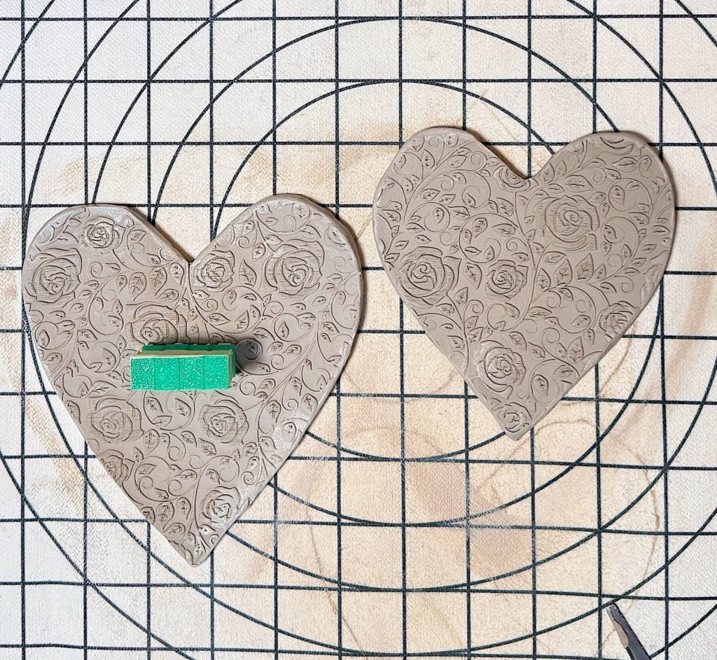 Two heart-shaped clay slabs with embossed floral patterns are placed on a gridded surface; one has a small green rectangular object on it.