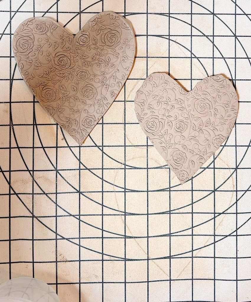 Two heart-shaped pieces of clay, decorated with a floral pattern, sit on a gridded work surface with circular guides.