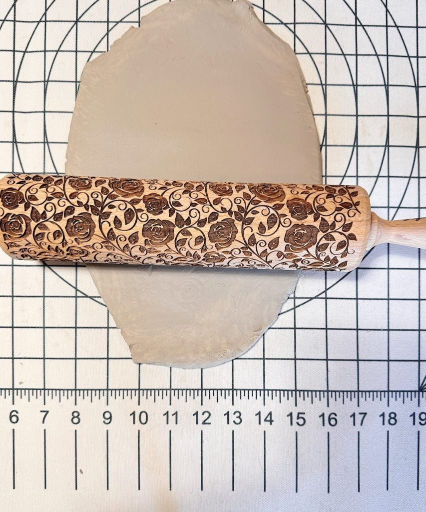 An embossed rolling pin with a floral pattern is being used to roll out a sheet of clay on a gridded surface with a ruler.