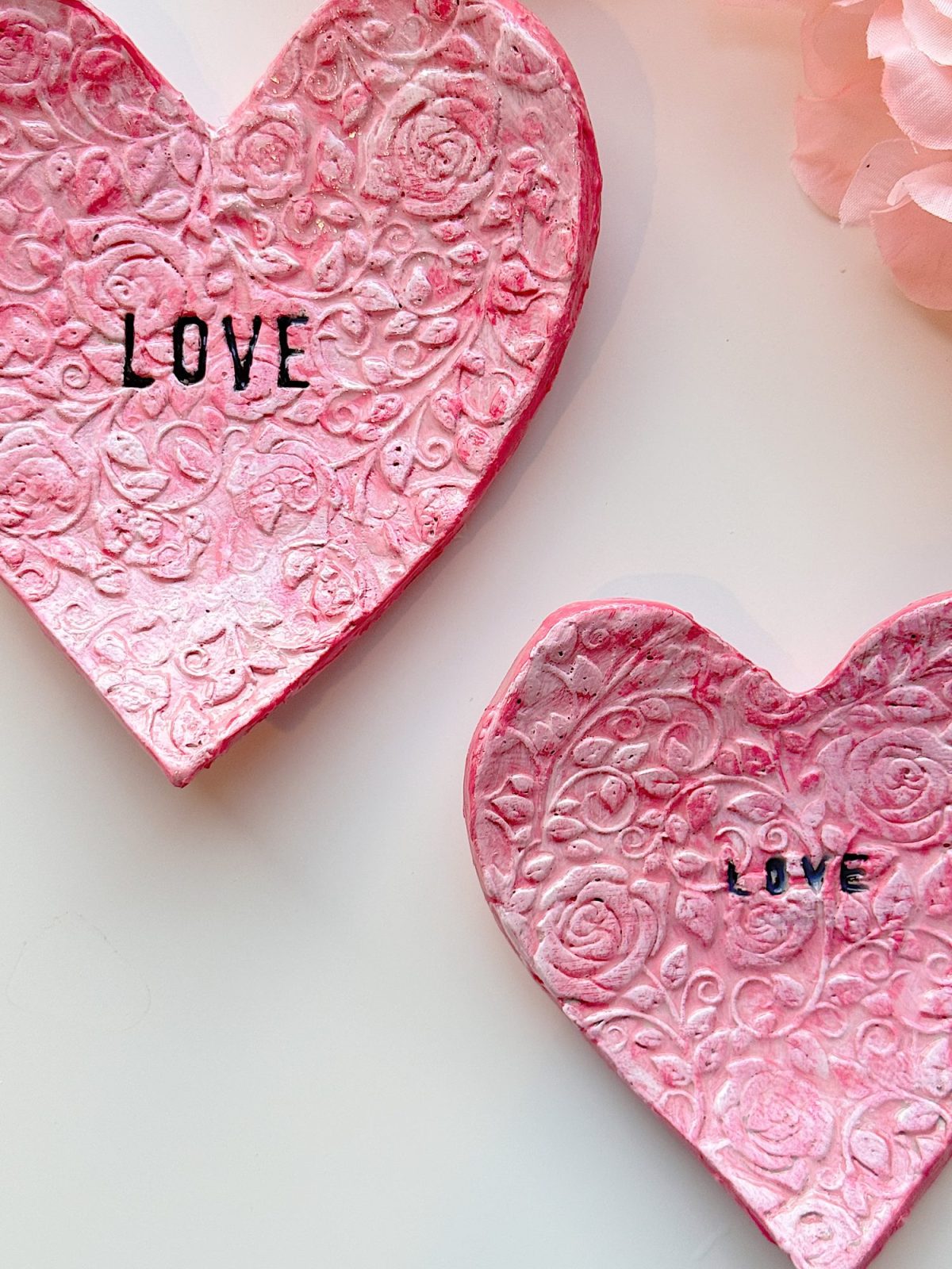 Two pink, heart-shaped clay dishes with embossed floral patterns and the word "LOVE" stamped in black letters on each, placed on a white surface.