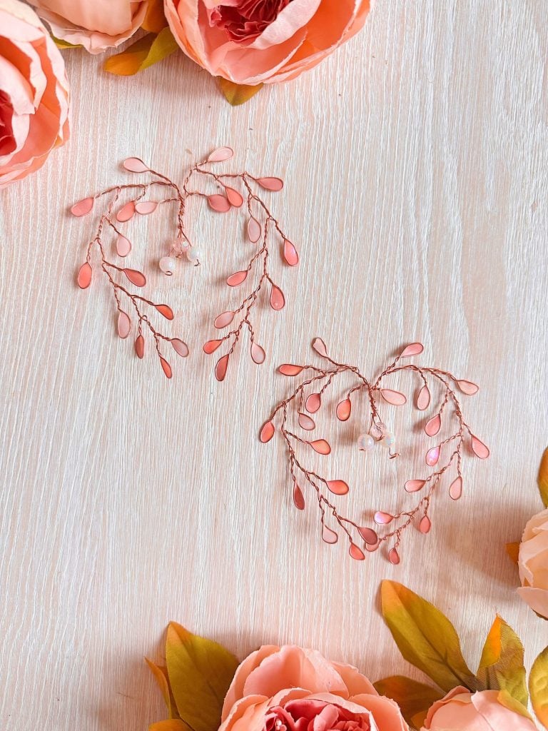 Two heart-shaped arrangements of pink and coral beads on wire are placed on a light wood surface, surrounded by peach-colored artificial flowers.