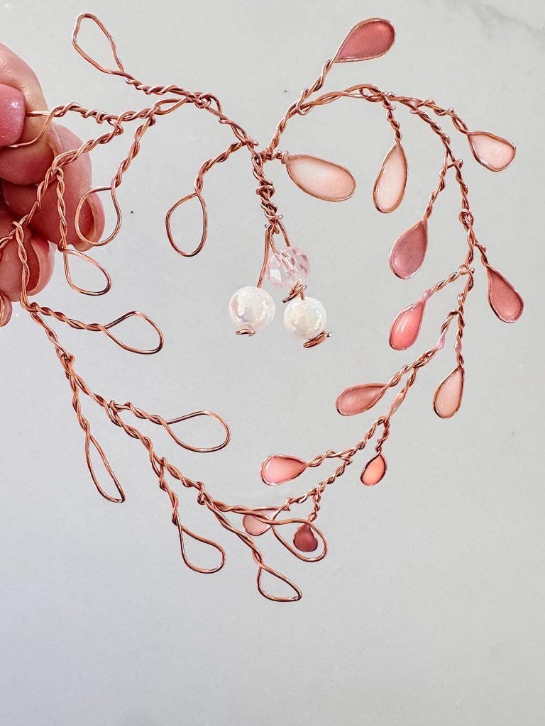 A hand holds a heart-shaped wire sculpture with pink and clear bead accents, featuring twisted wire branches and teardrop-shaped loops.