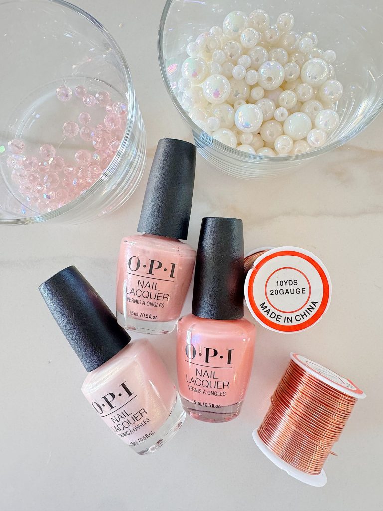 Three bottles of OPI nail lacquer, a spool of copper wire, and two bowls of assorted beads are arranged on a light surface.
