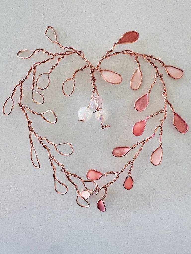 Heart-shaped arrangement of twisted copper wire branches with pink teardrop beads and three round white beads in the center, on a light surface.