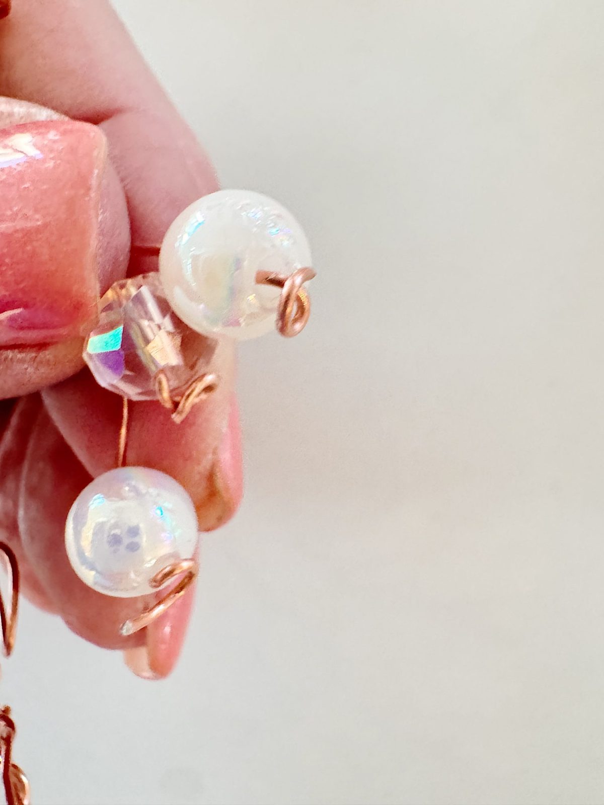 A hand holding a beaded ornament with iridescent white and clear beads, secured by copper wire against a plain background.