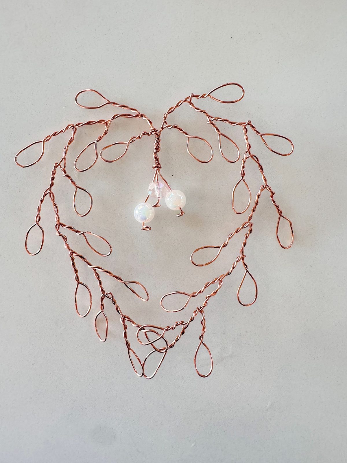 Twisted copper wire forms a heart-shaped tree design with three iridescent beads hanging in the center, set against a plain light background.
