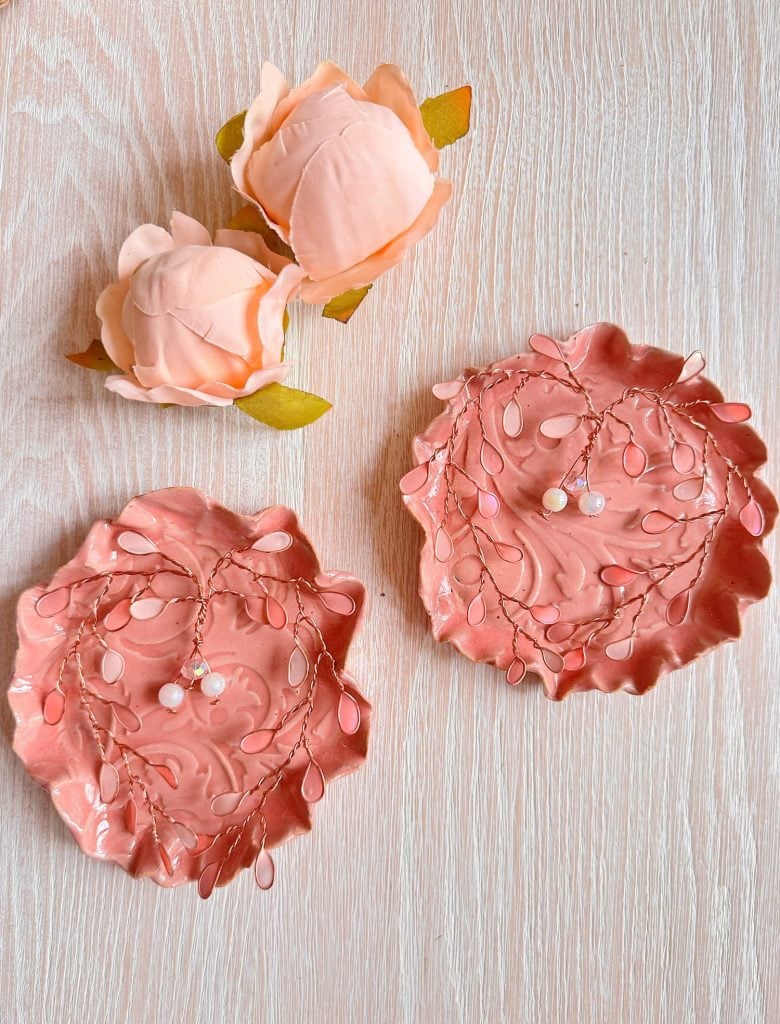 Two pink decorative dishes with leaf designs and beaded wire d&eacute;cor are placed on a light wood surface, alongside two artificial pink roses.