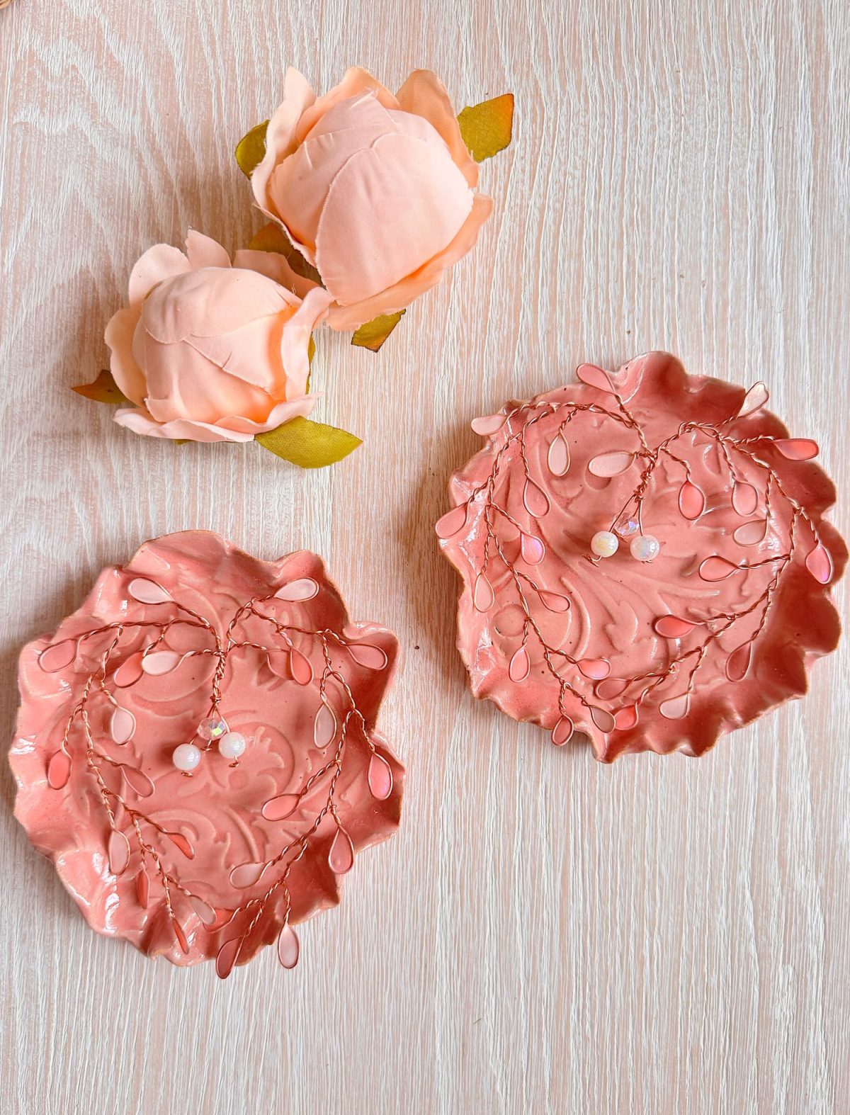 Two pink decorative dishes with leaf designs and beaded wire décor are placed on a light wood surface, alongside two artificial pink roses.