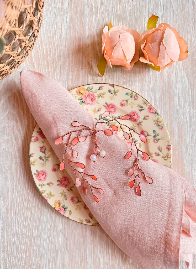 A floral-patterned plate with a pink napkin, a decorative pink and pearl branch, and two artificial peach roses on a light wood surface.