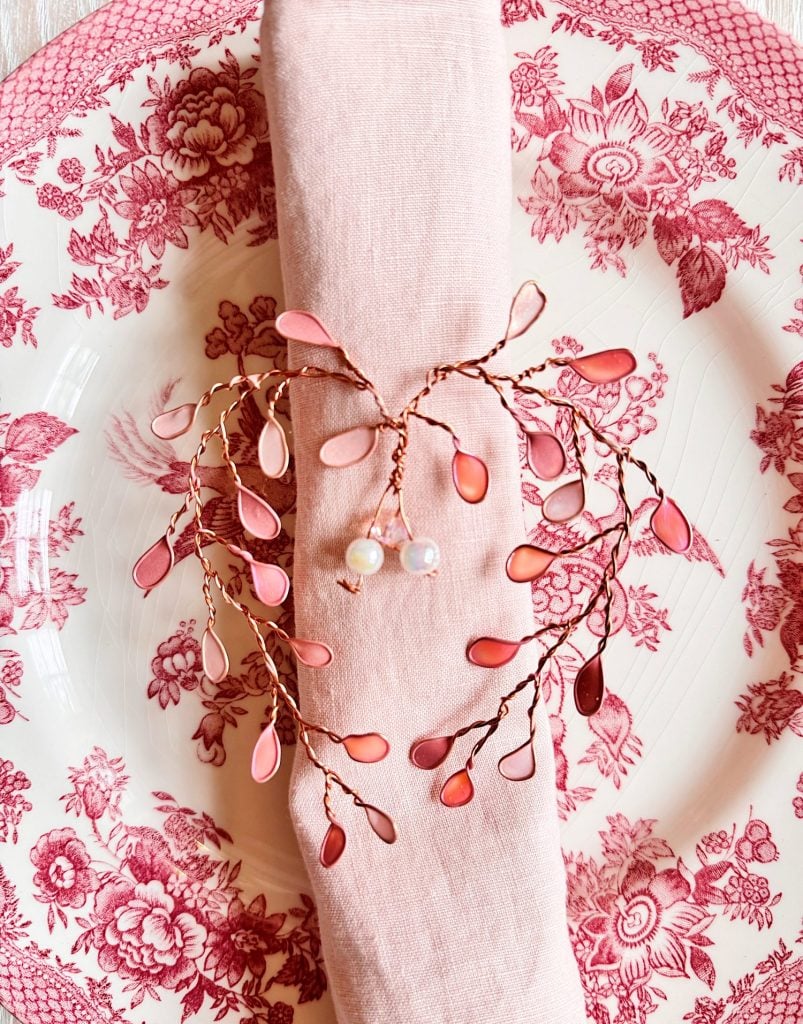 Pink napkin on a pink and white floral plate, held by a decorative napkin ring with pink leaf-shaped beads and two pearl-like beads.
