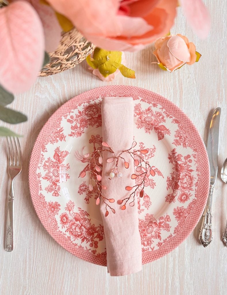 A pink and white floral plate with a pink napkin and decorative ring is set on a light wooden table with a fork, knife, spoon, and artificial flowers.