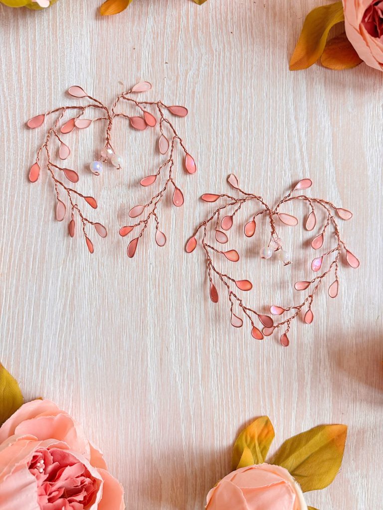 Two decorative, heart-shaped wire ornaments with pink beads are placed on a light wooden surface, surrounded by artificial pink flowers.