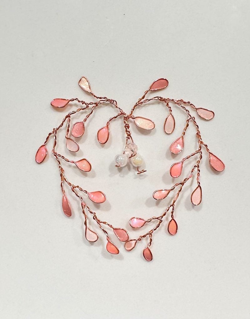 A decorative wire ornament with pink, leaf-shaped segments arranged in a heart shape, featuring three white bead accents in the center, on a plain light background.