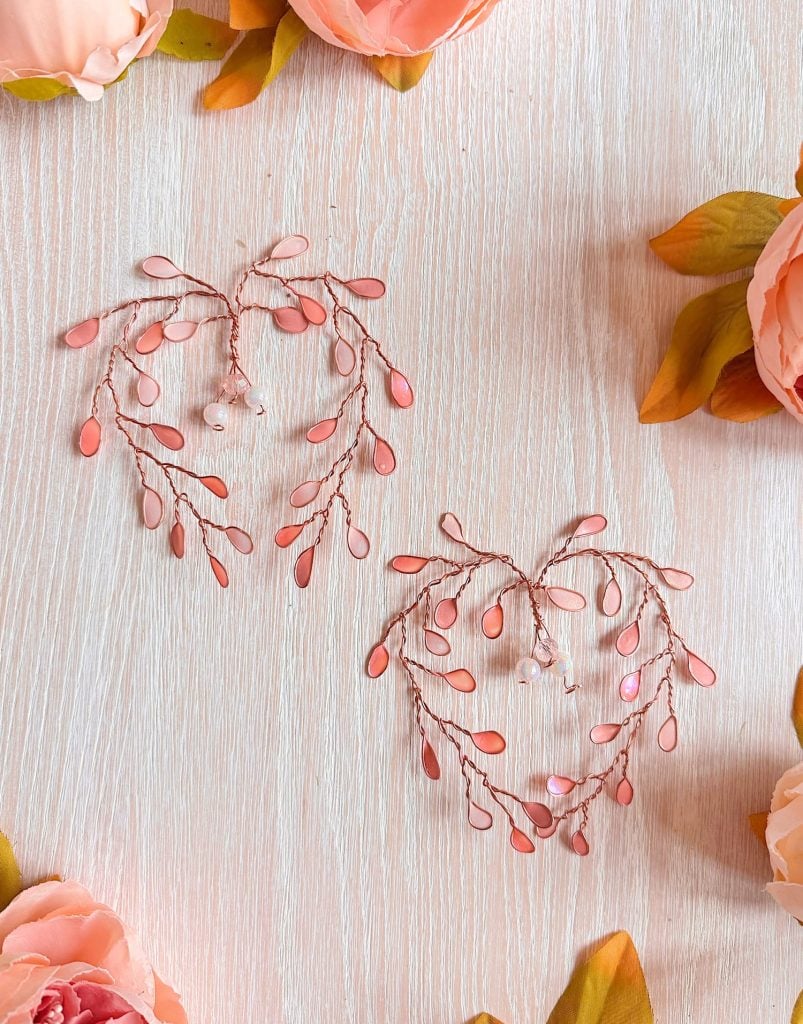 Two heart-shaped arrangements made of pink and peach beads with wire branches are placed on a light wooden surface, surrounded by artificial peach-colored flowers.
