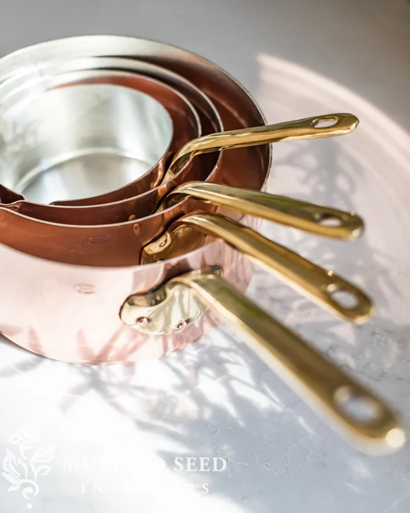 Four nested copper saucepans with gold-colored handles are arranged on a light surface, casting shadows.