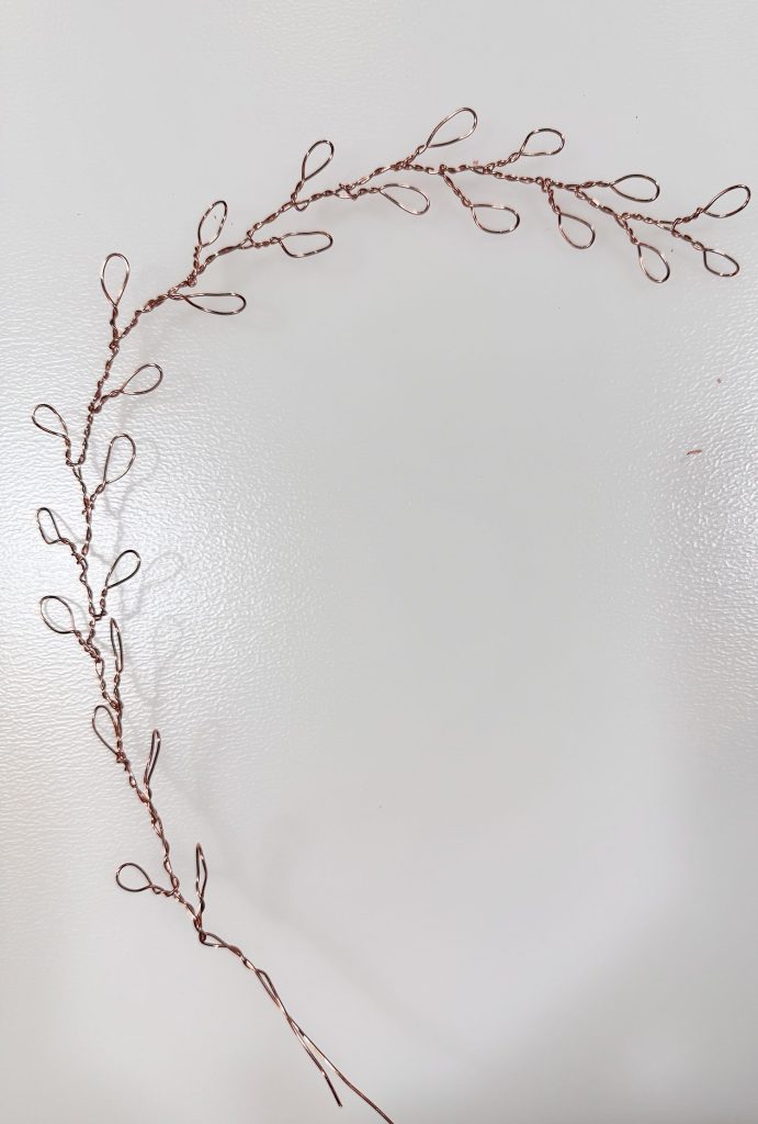 A handcrafted wire branch with looped wire leaves arranged in a curved, semi-circular shape on a white background.