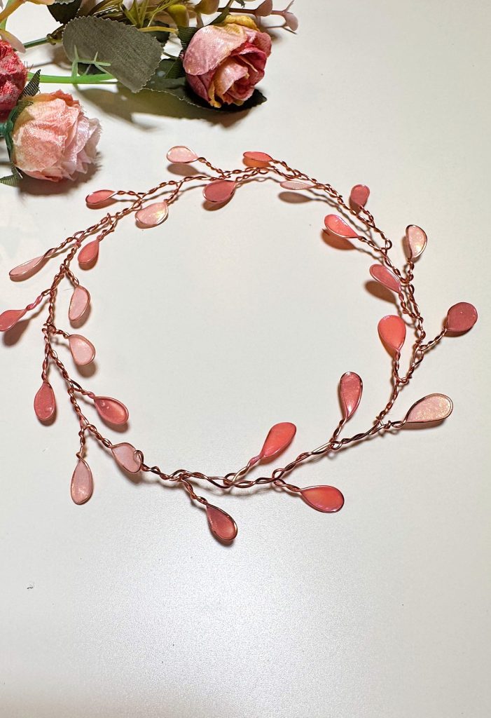A delicate pink floral headpiece with teardrop-shaped beads and wire, arranged in a circular shape on a white surface, with artificial flowers in the corner.
