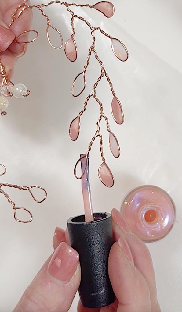A hand holds a nail polish brush and applies pink nail polish to small wire loops shaped like leaves on a decorative heart wreath branch.