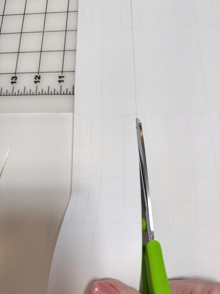 A hand holding green scissors cuts along a faint pencil line on white paper with a gridded cutting mat visible in the background.