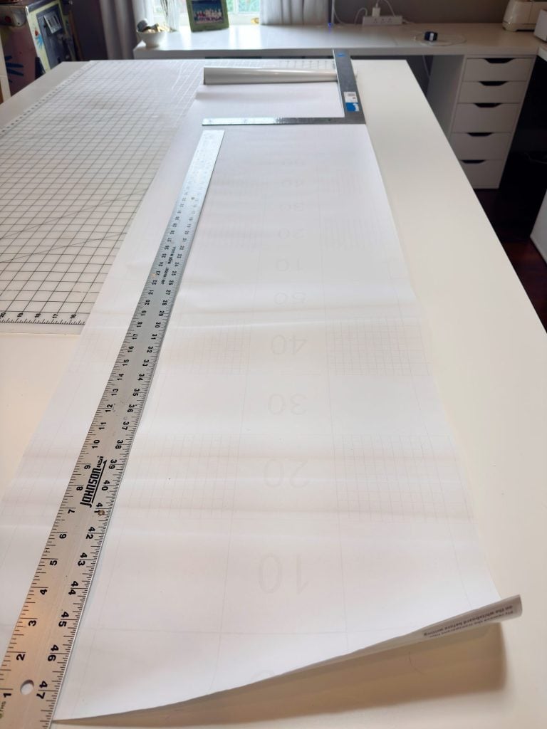 A long sheet of paper with numbers printed on it is laid out on a white table, next to a metal ruler and a large cutting mat in a well-lit workspace.