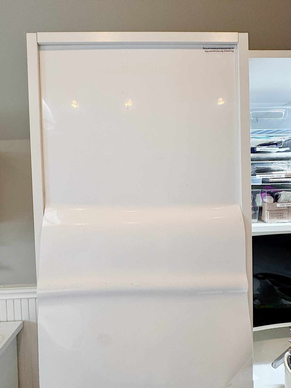 A white flip chart with a wrinkled sheet of blank paper is displayed on an easel in a room with storage shelves in the background.