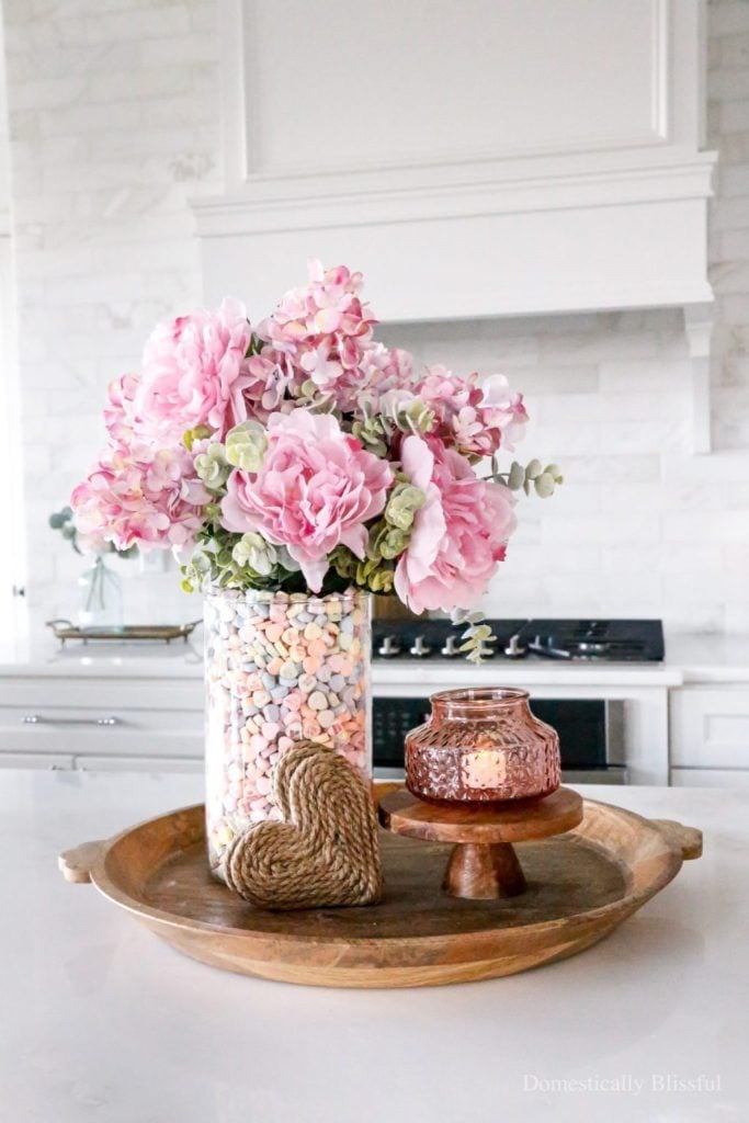 A glass vase filled with pastel candy hearts and pink flowers sits on a wooden tray with a twine-wrapped decorative heart and a small pink glass candle holder in a white kitchen.