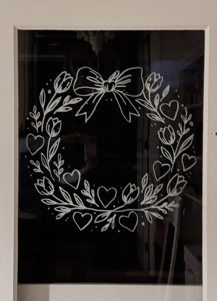 A white line drawing of a wreath with leaves, hearts, and a bow is displayed on a window or glass door.
