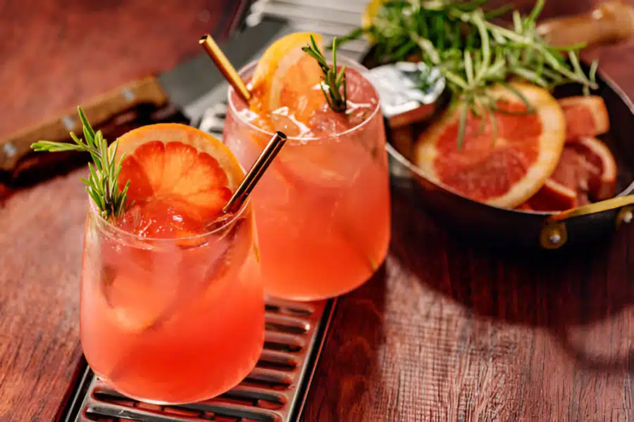 Two glasses of pink grapefruit cocktail with metal straws, garnished with grapefruit slices and rosemary, on a wooden surface next to a pan of sliced grapefruit and herbs.