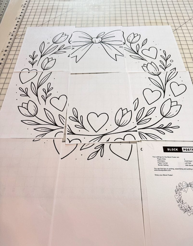 Six sheets of paper form a large black-and-white line drawing of a wreath with flowers, leaves, hearts, and a bow, laid out on a gridded cutting mat.