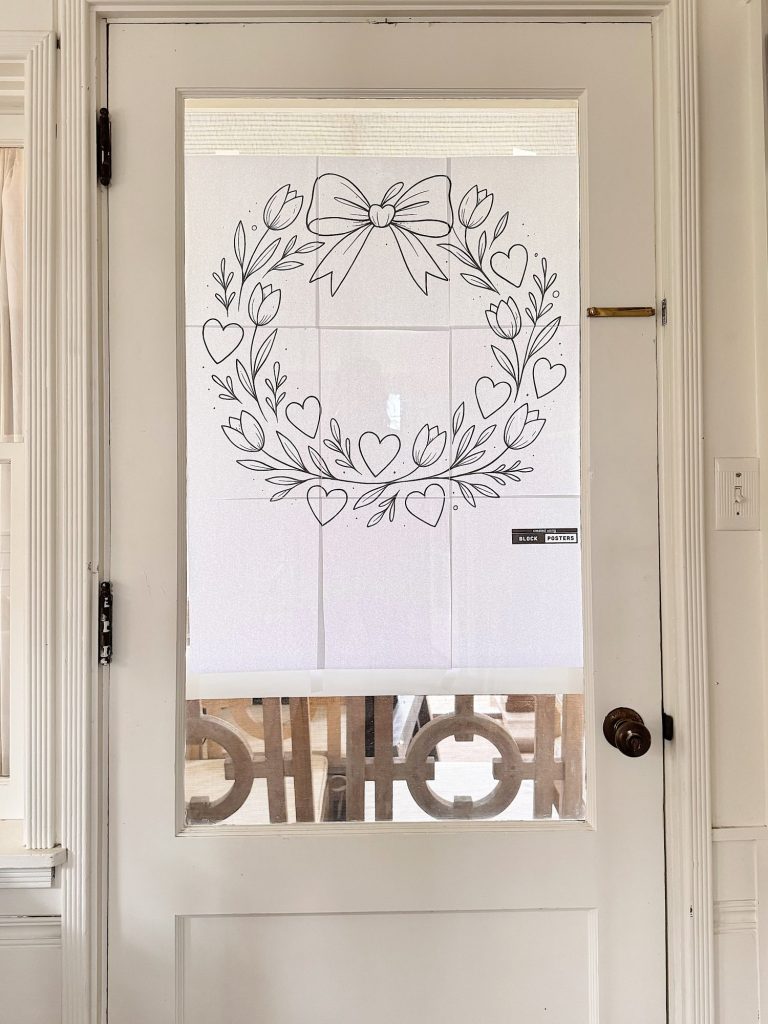 A white door with a glass window covered by a sheer curtain featuring a black outline drawing of a wreath with hearts and a bow.