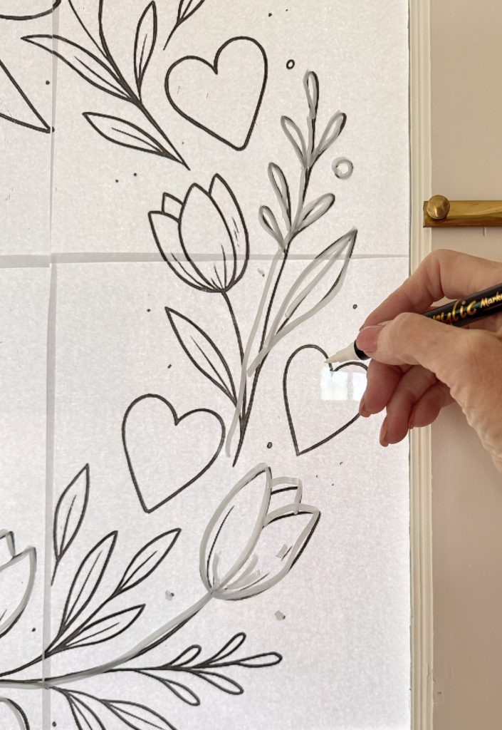 A hand traces black floral and heart designs on a white sheet using a clear adhesive or pen, with a brass door handle visible on the right side.