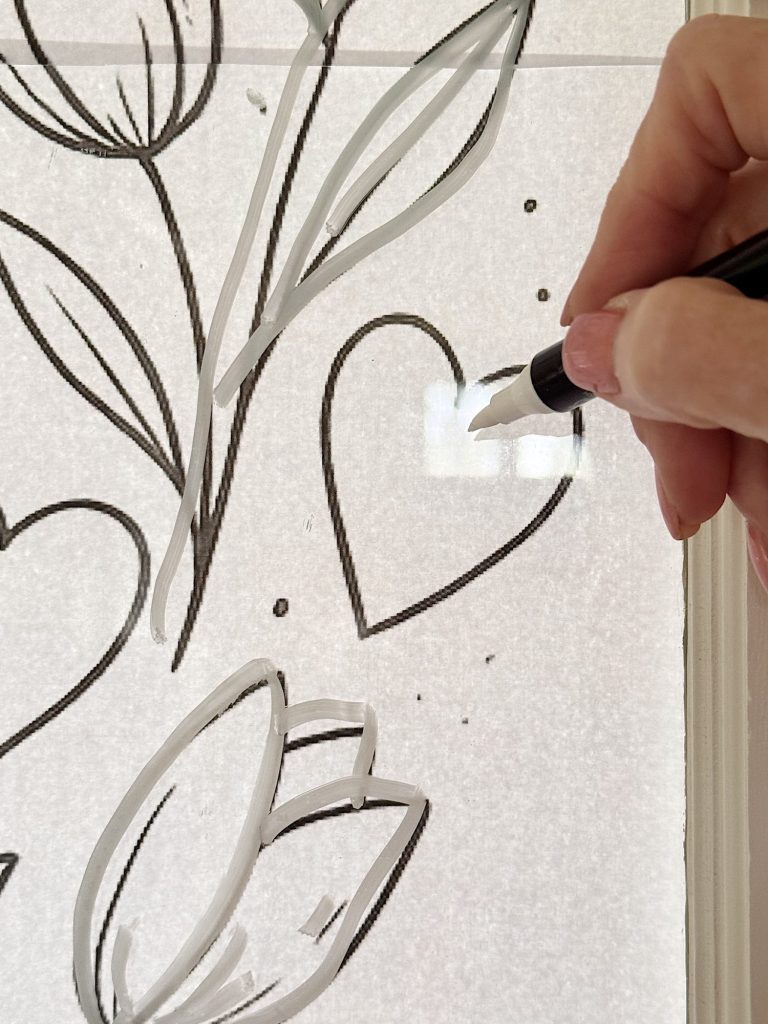 A hand uses a black marker to draw a heart and flowers on a transparent surface.