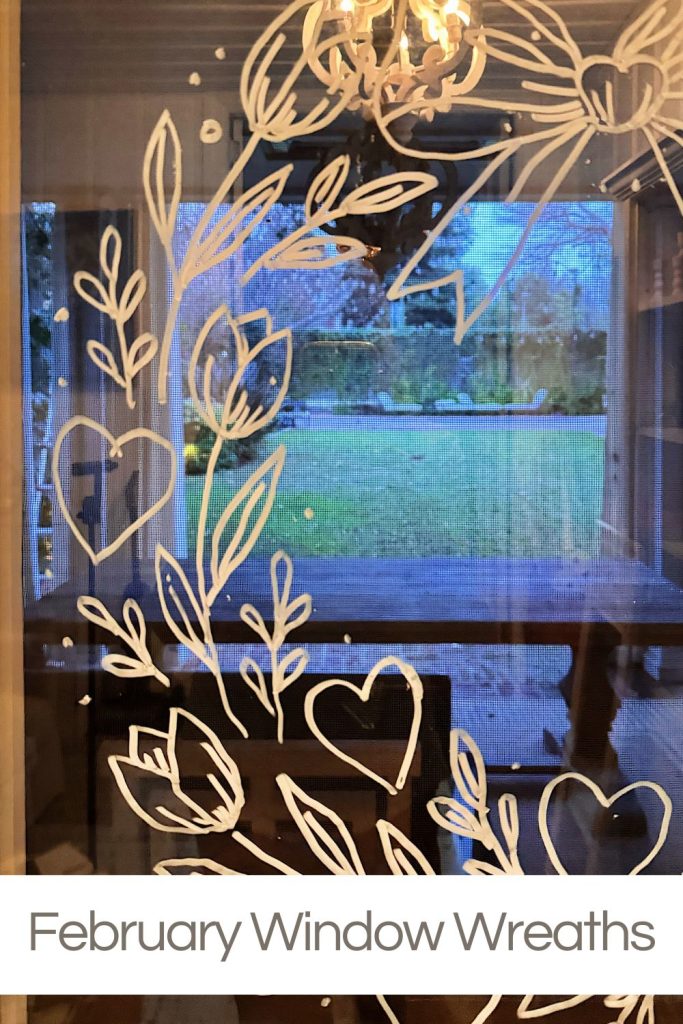 White hearts and leafy wreath designs drawn on a window, with a view of a green lawn and trees outside. Text at the bottom reads "February Window Wreaths.