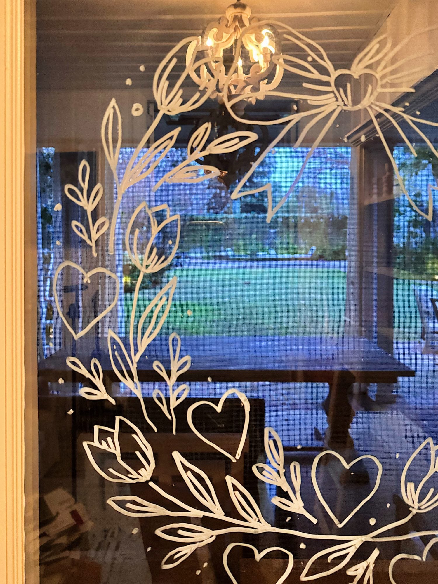 White floral and heart designs are drawn on a glass door, with a view of a wooden table, chairs, and a grassy backyard visible through the glass.