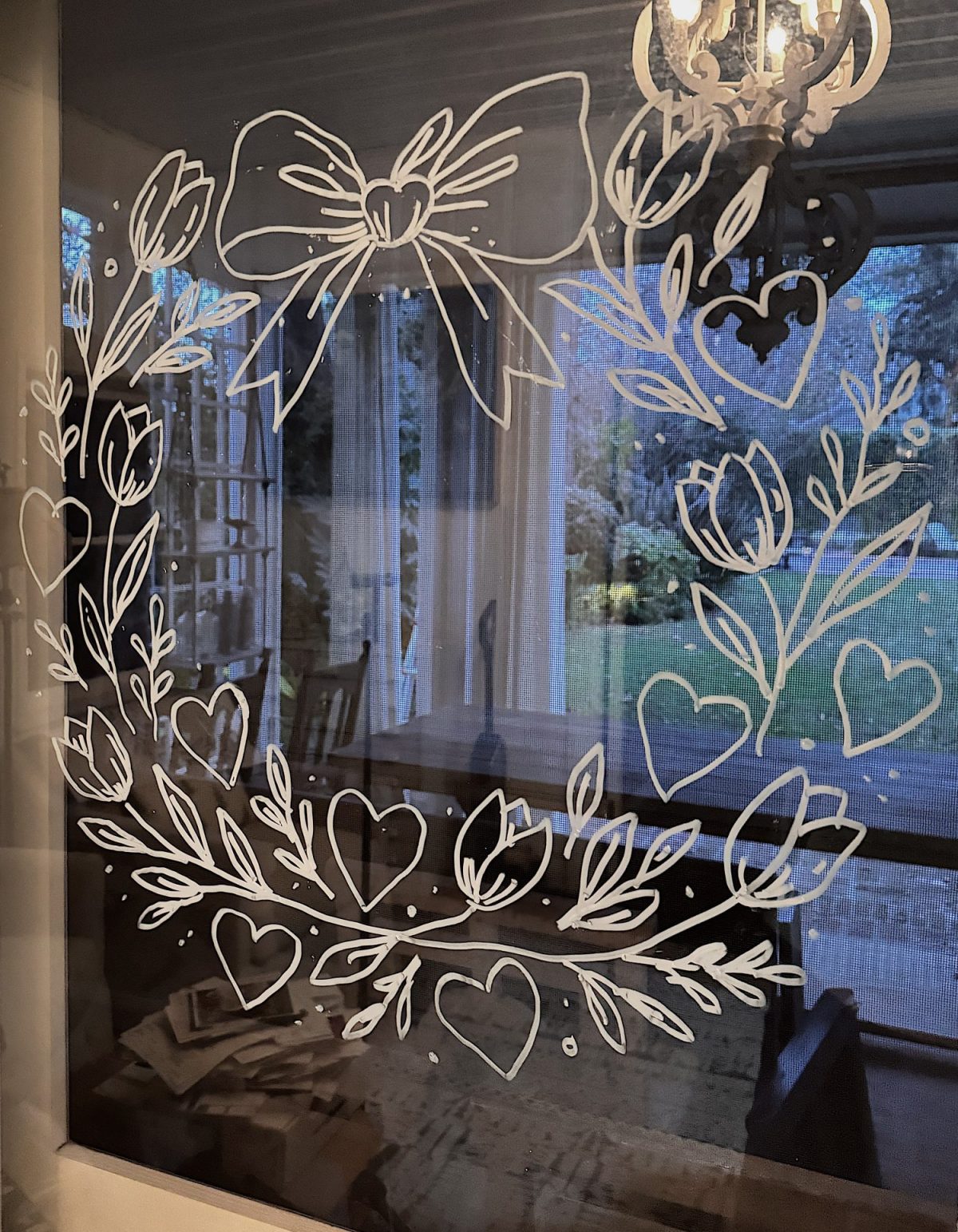 White window drawing of a large bow, leaves, tulips, and hearts, with an indoor dining area and garden visible through the glass.