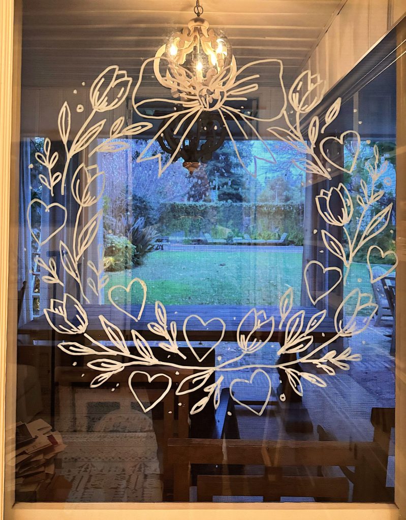 White marker drawing of a heart-themed wreath with leaves and dots on a window, showing a green yard and trees outside, and a chandelier reflecting in the glass.