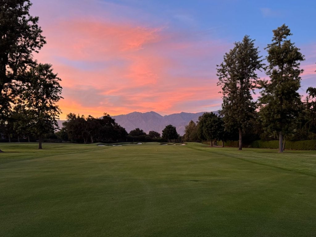 Golf course fairway at sunset with mountains in the background and trees lining both sides of the green under a pink and blue sky.