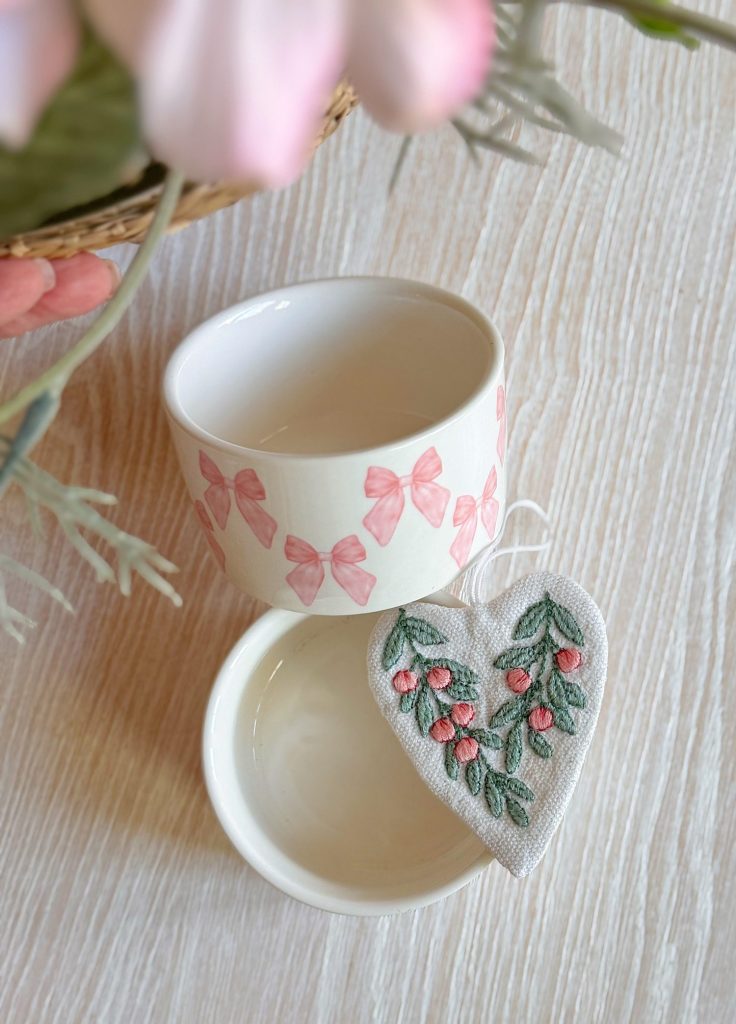 A white ceramic bowl with pink bow designs, another plain white bowl, and a heart-shaped fabric ornament with embroidered flowers on a light wooden surface.