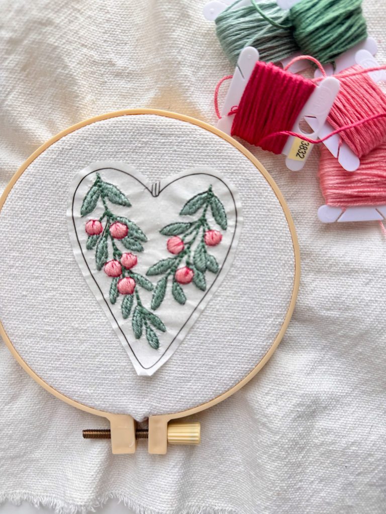 An embroidery hoop with a heart-shaped floral design in pink and green thread, next to pink and green embroidery floss on a textured white fabric background.