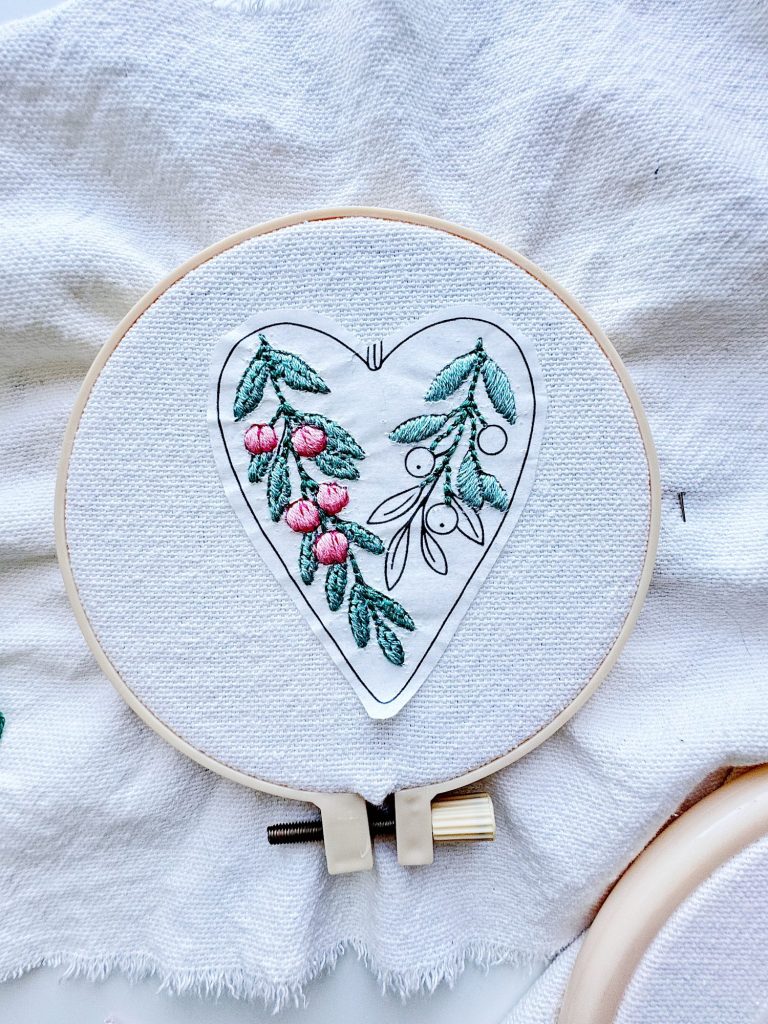 Embroidery hoop with white fabric showing a heart-shaped design featuring green leaves and pink berries stitched in thread.